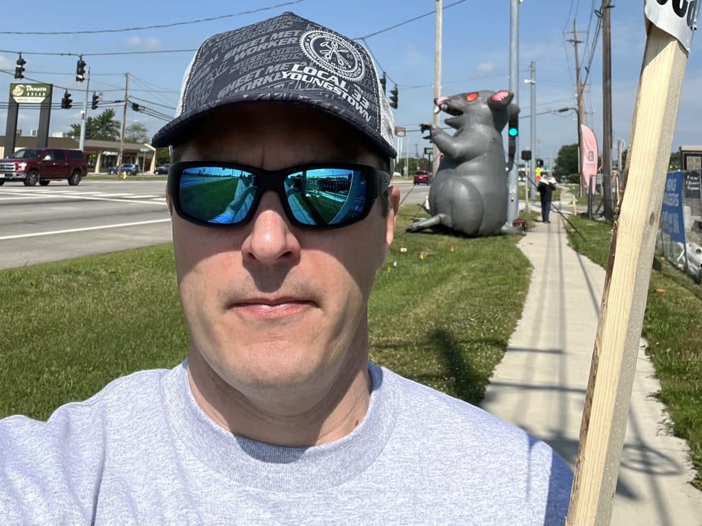 SMART Education Department Director Eli Baccus on the picket line during his time as an organizer with Local 33, pictured wearing a Local 33 hat with an inflatable Scabby the Rat behidn him.